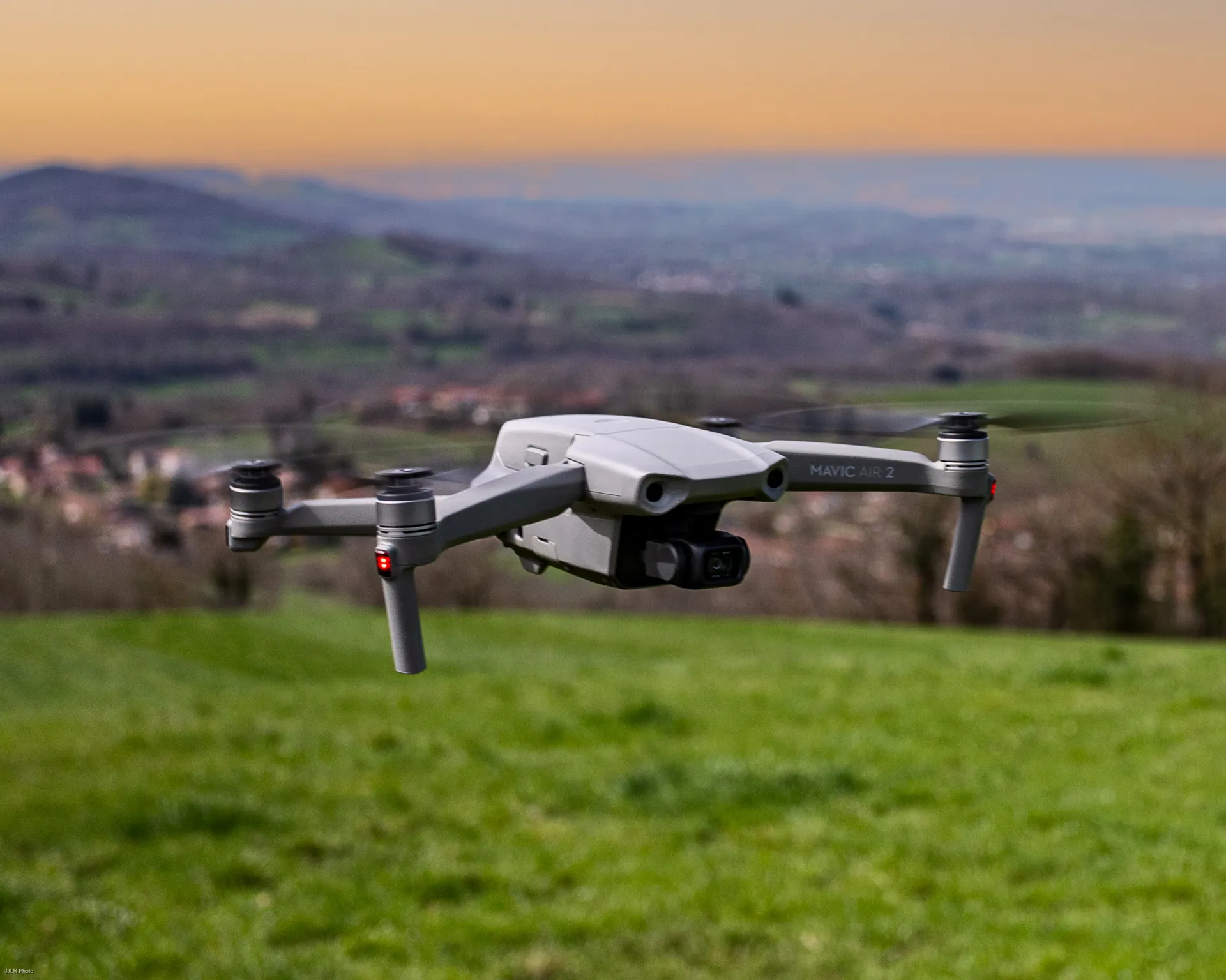 Un drone gris en vol stationnaire au-dessus d'un pré, la campagne et les collines derrière, au soleil couchant.