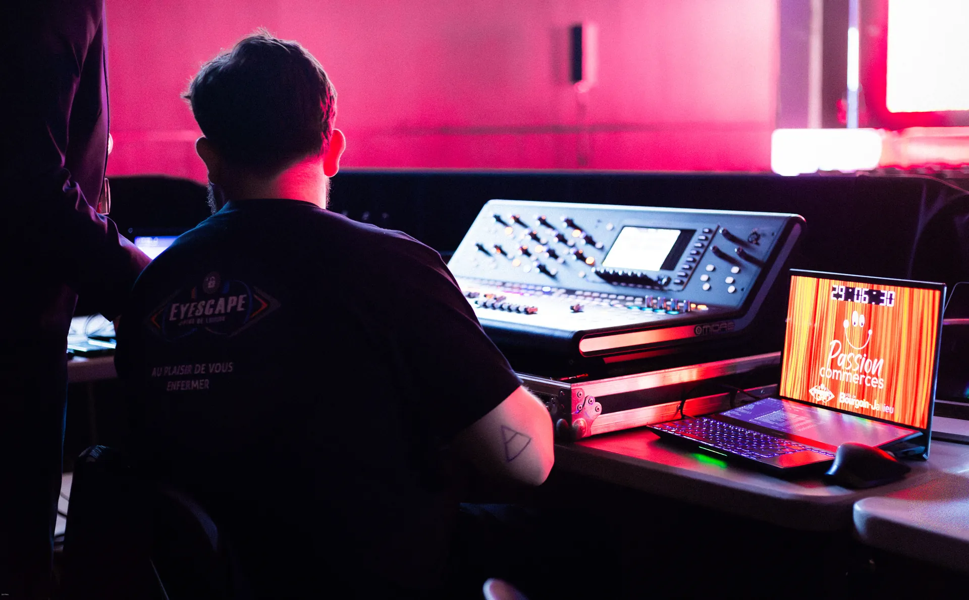 Un technicien de dos devant une console de mixage, dans une lumière rouge. Sur l'écran d'un ordinateur à côté, un compte à rebours et les mots Passion commerces.